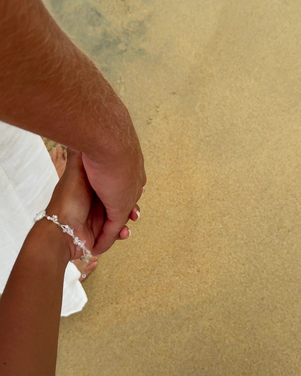 Two hands held together over pale sand, one wearing a beaded bracelet.