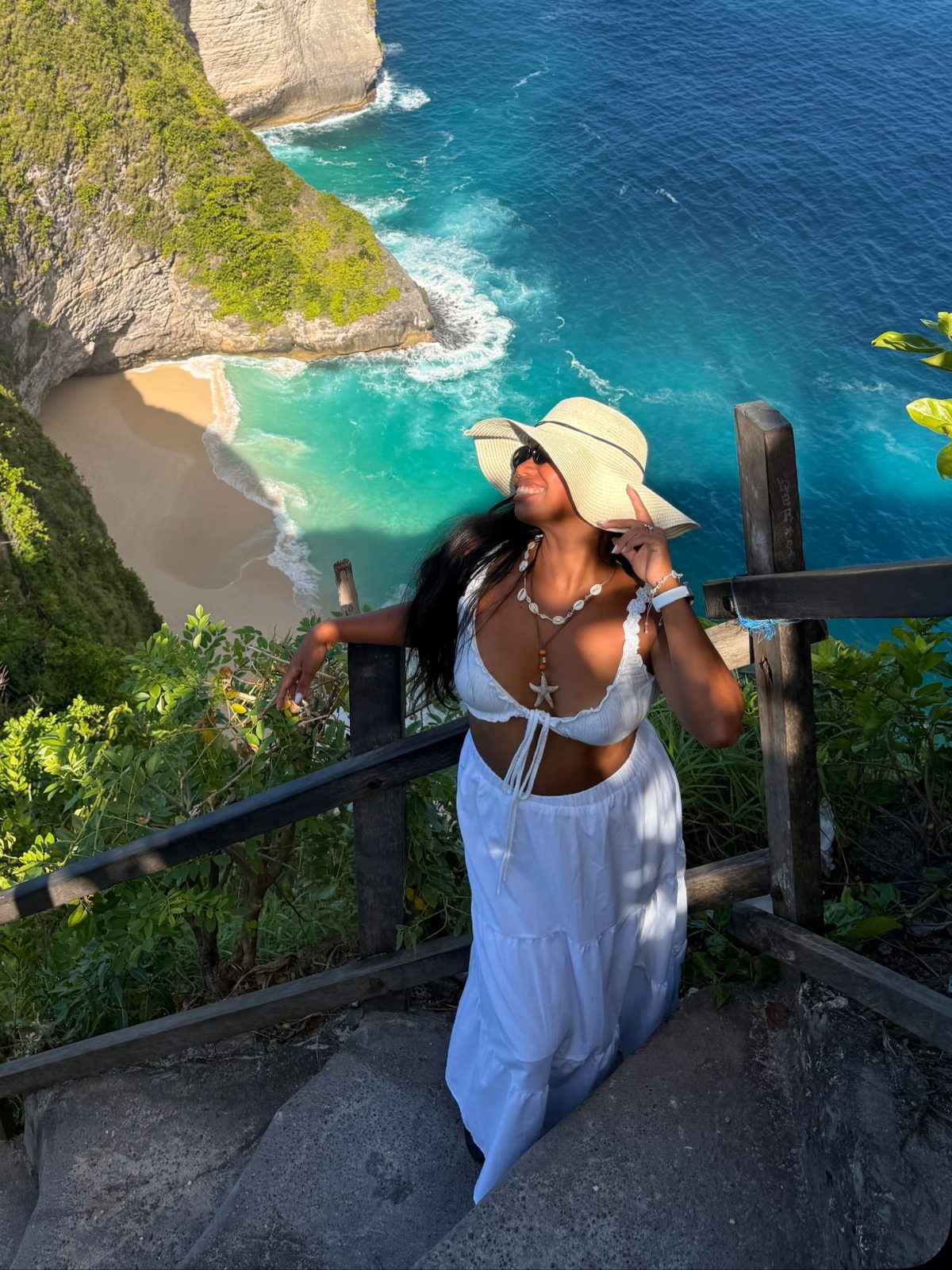 Vivian Dolor standing on a cliff above a turquoise bay in Nusa Penida, wearing a white top and sun hat.